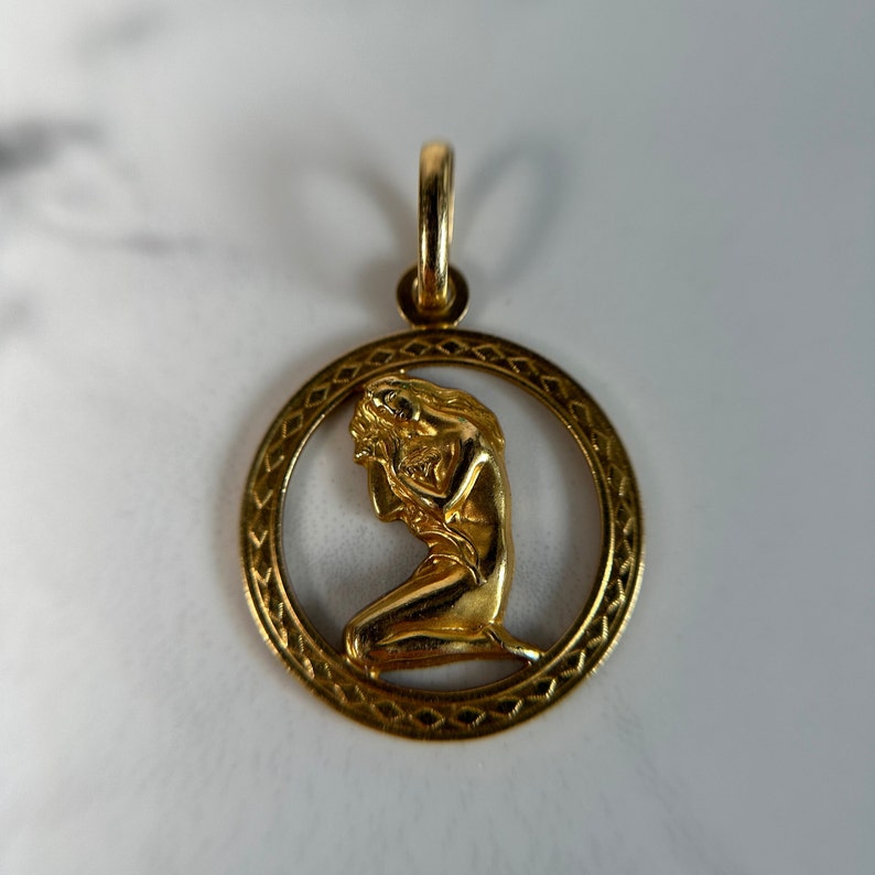 May include: Gold pendant featuring a detailed, three-dimensional figure of a woman kneeling inside a circular frame with a geometric design.