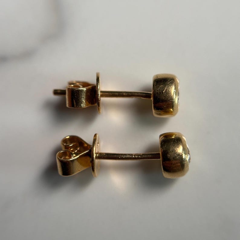 May include: A pair of gold stud earrings. Each earring features a small, round, clear gemstone set in a gold bezel. The earrings have a post and butterfly backing. The earrings are displayed on a light-colored surface.