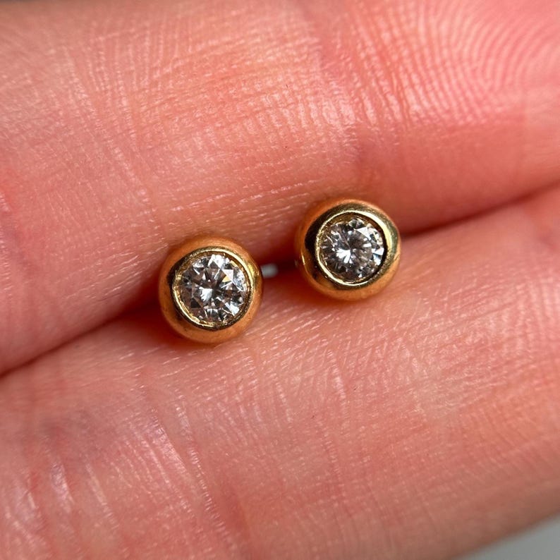 May include: A pair of gold stud earrings, each featuring a round, clear gemstone set in a bezel. The earrings have a simple, classic design with a polished gold finish, suitable for everyday wear.