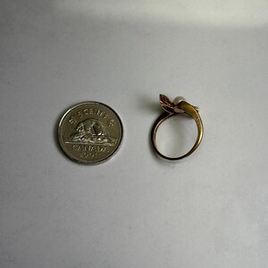 May include: A gold-toned ring with a pearl and a floral design, next to a Canadian 5-cent coin. The ring features a delicate, curved band and a small, round pearl. The coin is silver and shows a bear.