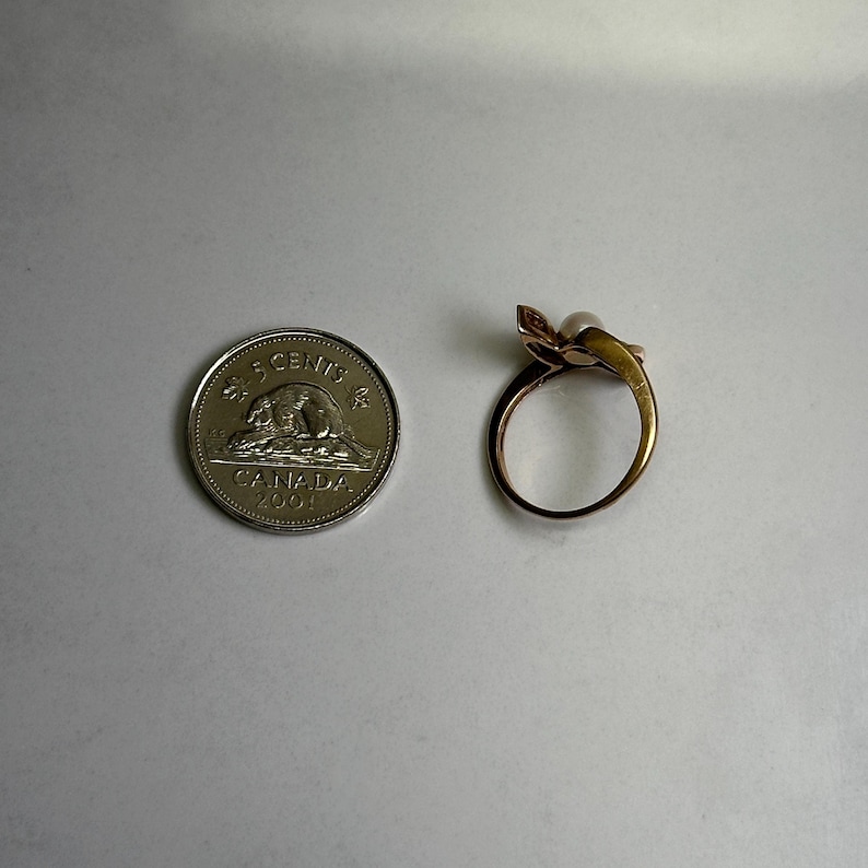 May include: A gold-toned ring with a pearl and a floral design, next to a Canadian 5-cent coin. The ring features a delicate, curved band and a small, round pearl. The coin is silver and shows a bear.