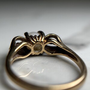 Puede incluir: Un anillo de oro con una perla blanca engastada en un engaste decorativo. El anillo se ve desde abajo, mostrando la parte inferior de la perla y los intrincados detalles del engaste.