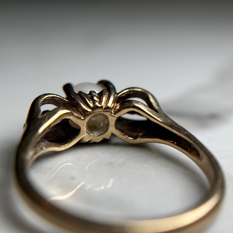 Puede incluir: Un anillo de oro con una perla blanca engastada en un engaste decorativo. El anillo se ve desde abajo, mostrando la parte inferior de la perla y los intrincados detalles del engaste.