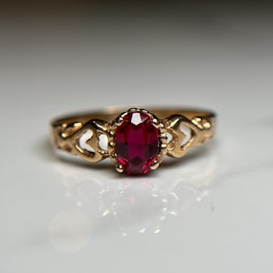 May include: A gold ring featuring a red oval-shaped gemstone set in a heart-shaped design.