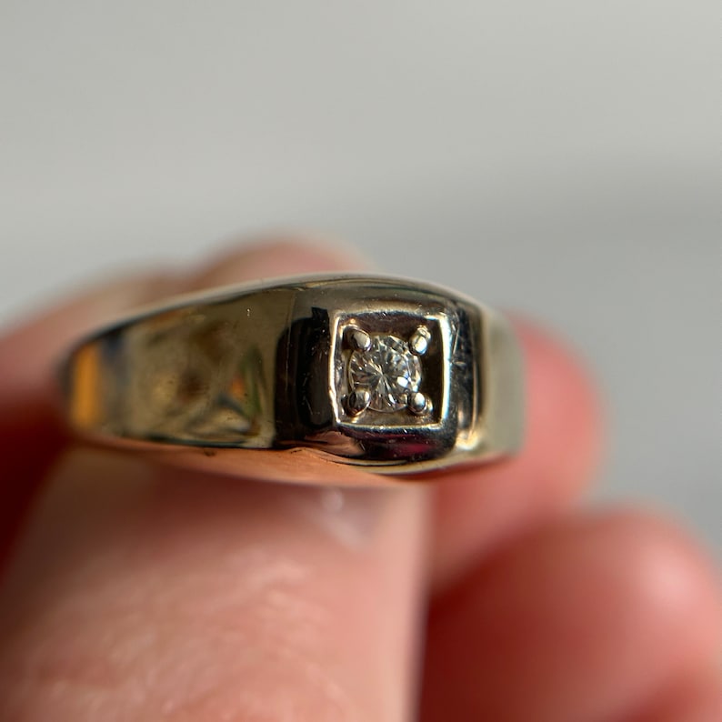 May include: A gold band ring with a single, clear diamond set in a square bezel.