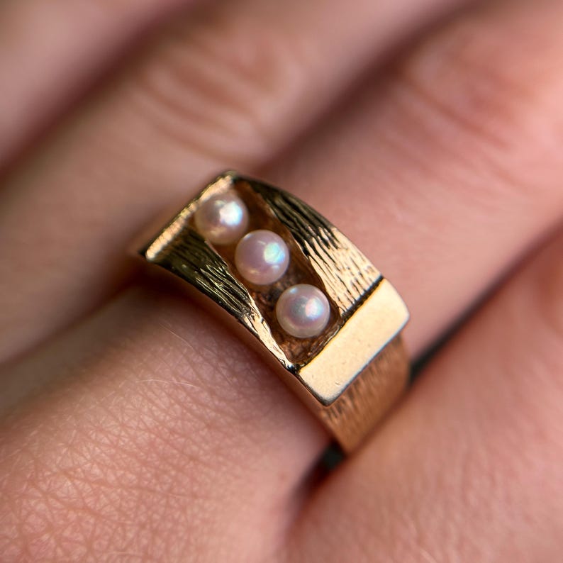 May include: A gold-toned ring featuring three small, round pearls set in a rectangular design. The ring has a textured band and a polished finish, showcasing a classic and elegant style.