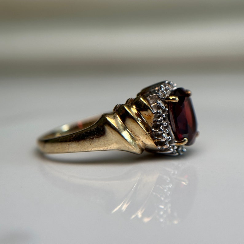 May include: A gold ring featuring a large, dark red gemstone surrounded by smaller, clear gemstones.