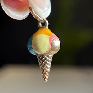 May include: A silver ice cream cone charm with three scoops of ice cream in yellow, green, pink, and blue. The charm is held up by a silver chain. The ice cream cone has a textured design.