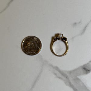 Puede incluir: Un anillo de oro con una sola perla en el centro. El anillo est&aacute; engastado con peque&ntilde;os diamantes a cada lado de la perla.