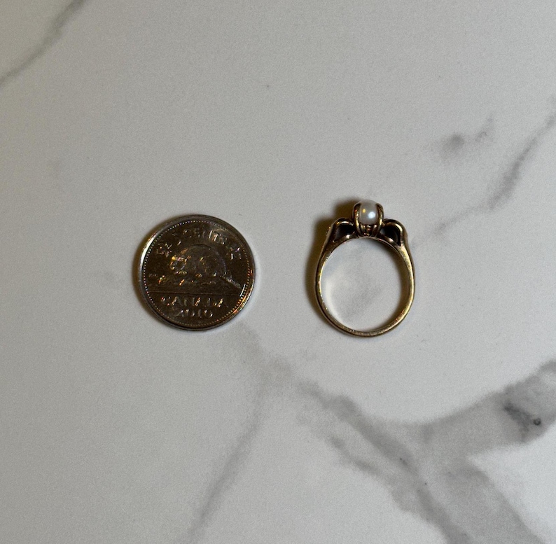 Puede incluir: Un anillo de oro con una sola perla en el centro. El anillo est&aacute; engastado con peque&ntilde;os diamantes a cada lado de la perla.