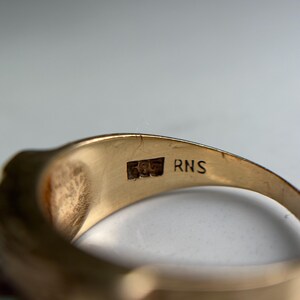 May include: Close-up of a gold ring with a smooth, polished surface. The ring has a hallmark with the numbers "585" and the letters "RNS" engraved on the inside band. The ring is a warm, yellow color.