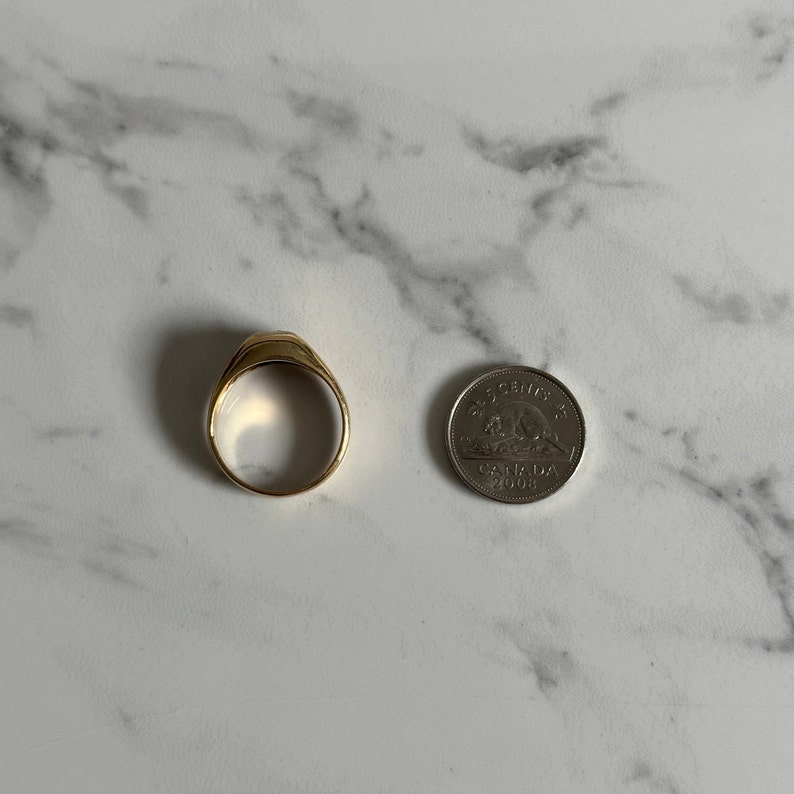 May include: A gold signet ring with a flat top, shown next to a Canadian 25-cent coin for size comparison.
