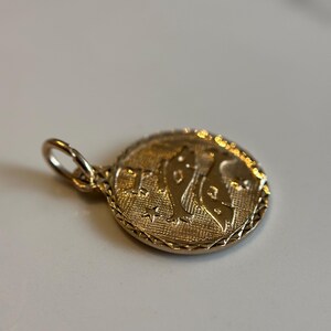 Puede incluir: Un colgante dorado con una superficie texturizada. El colgante presenta un dise&ntilde;o en relieve de una figura y estrellas dentro de un marco circular. Se adjunta un peque&ntilde;o lazo para una cadena.