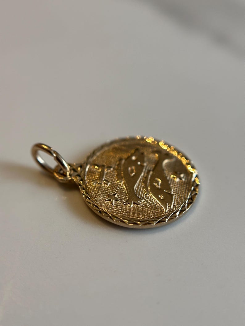 Puede incluir: Un colgante dorado con una superficie texturizada. El colgante presenta un dise&ntilde;o en relieve de una figura y estrellas dentro de un marco circular. Se adjunta un peque&ntilde;o lazo para una cadena.