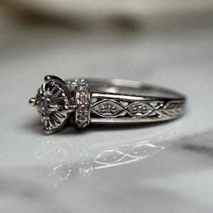 May include: A silver engagement ring with a delicate filigree design and a round center stone. The band is adorned with small diamonds.