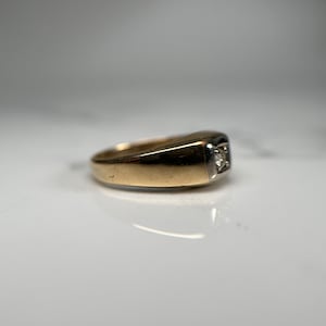 May include: A gold ring with a single diamond set in a square shape. The ring is made of two-toned metal, with a gold band and a white metal setting.