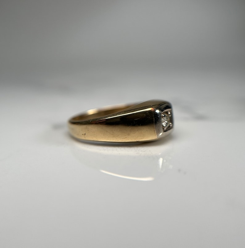 May include: A gold ring with a single diamond set in a square shape. The ring is made of two-toned metal, with a gold band and a white metal setting.