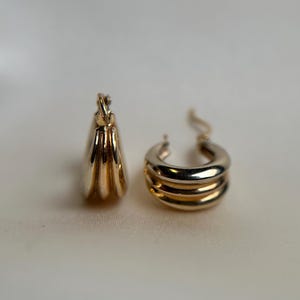 May include: A pair of gold hoop earrings with a triple-layered design. The earrings are made of two different shades of gold, creating a unique and stylish look.