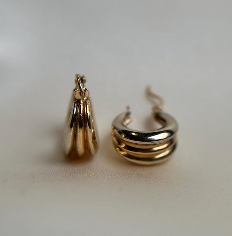 May include: A pair of gold hoop earrings with a triple-layered design. The earrings are made of two different shades of gold, creating a unique and stylish look.