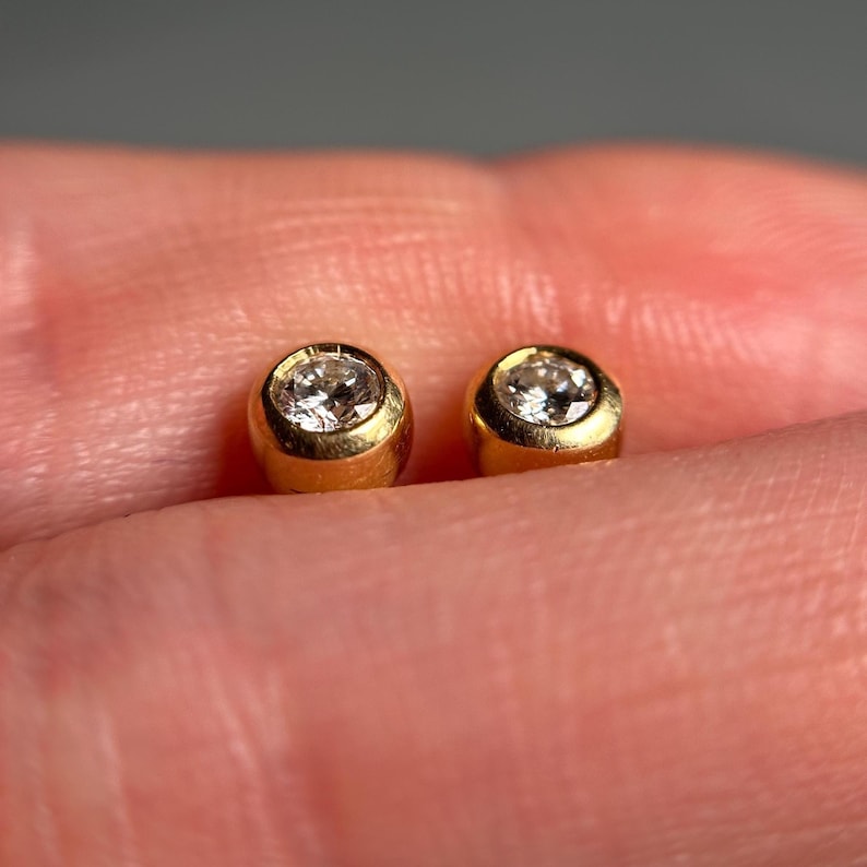 May include: Two small, round gold earrings with a single, clear gemstone set in each. The earrings are shown against a blurred background, highlighting the jewelry's design and sparkle. The earrings are a classic jewelry item.