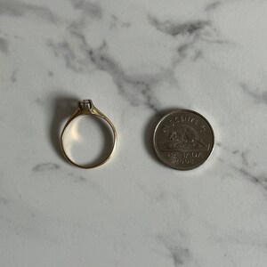 May include: A gold ring with a single clear stone set in the center. The ring is shown next to a Canadian 5-cent coin for size comparison.