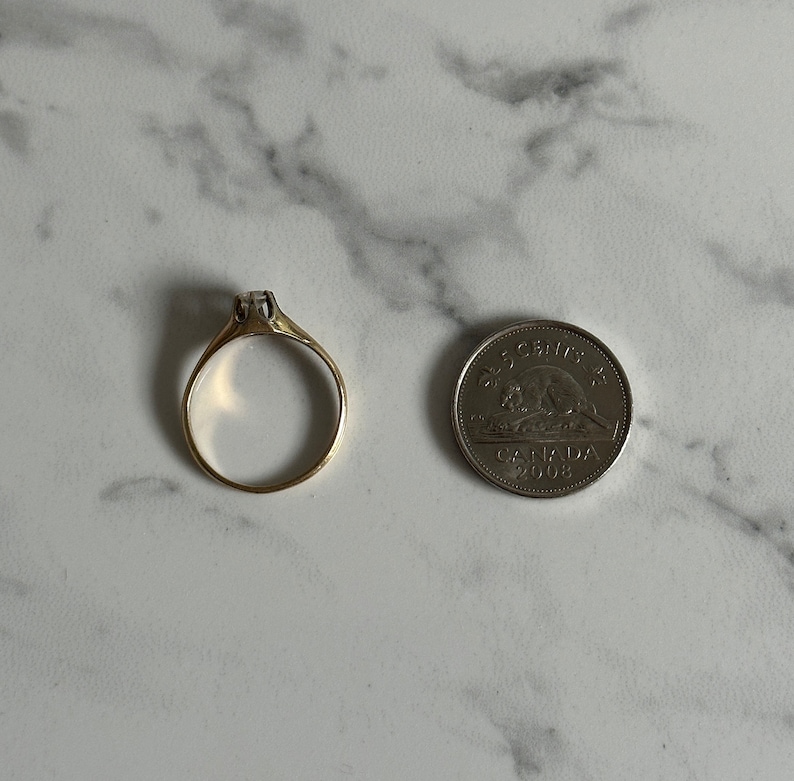 May include: A gold ring with a single clear stone set in the center. The ring is shown next to a Canadian 5-cent coin for size comparison.