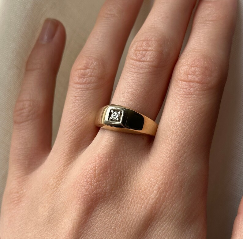 May include: A gold signet ring with a single, small, clear diamond set in the center.