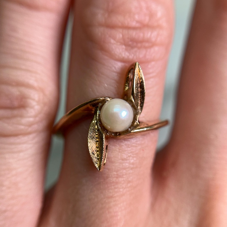 May include: A gold-toned ring featuring a central, round pearl. The ring's design incorporates stylized leaf accents that curve around the pearl, creating a floral or nature-inspired aesthetic. The ring is shown on a finger.