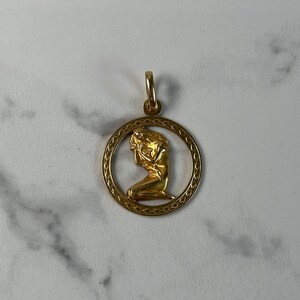 May include: Gold pendant featuring a detailed depiction of a woman in a kneeling pose, framed by a circular design with a decorative border.