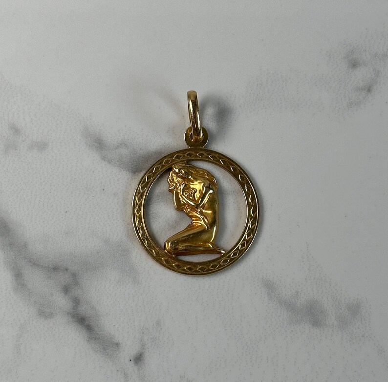 May include: Gold pendant featuring a detailed depiction of a woman in a kneeling pose, framed by a circular design with a decorative border.