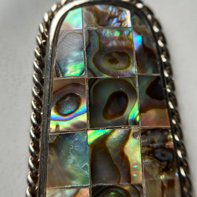 May include: A silver pendant with a rectangular abalone shell inlay. The shell is cut into small squares and arranged in a grid pattern. The shell has iridescent colors of green, blue, and brown.