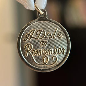May include: Gold-coloured circular pendant with the inscription "A Date To Remember" in elegant script. The pendant has a textured background and a loop for a chain. The text is raised and in a contrasting gold colour.