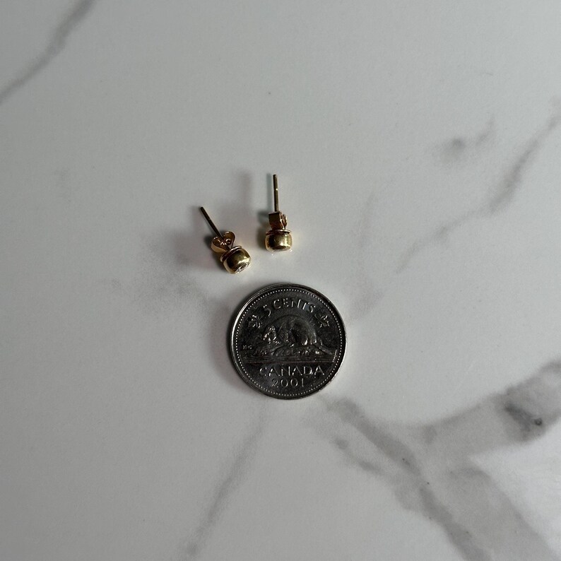 May include: A pair of small, gold-colored stud earrings with a round design. The earrings are displayed next to a Canadian five-cent coin, providing a size reference. The coin features a bear and the text "CANADA 2001".