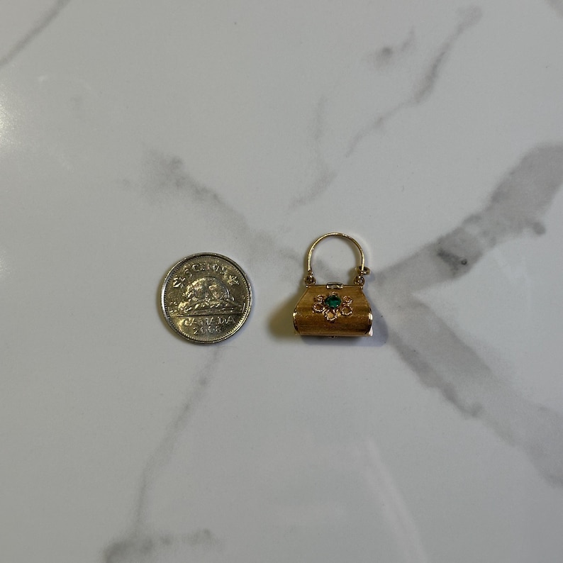 May include: A small, gold-colored purse charm with a green gemstone in the center of a floral design. The charm has a handle and a clasp. A Canadian coin is next to the charm.