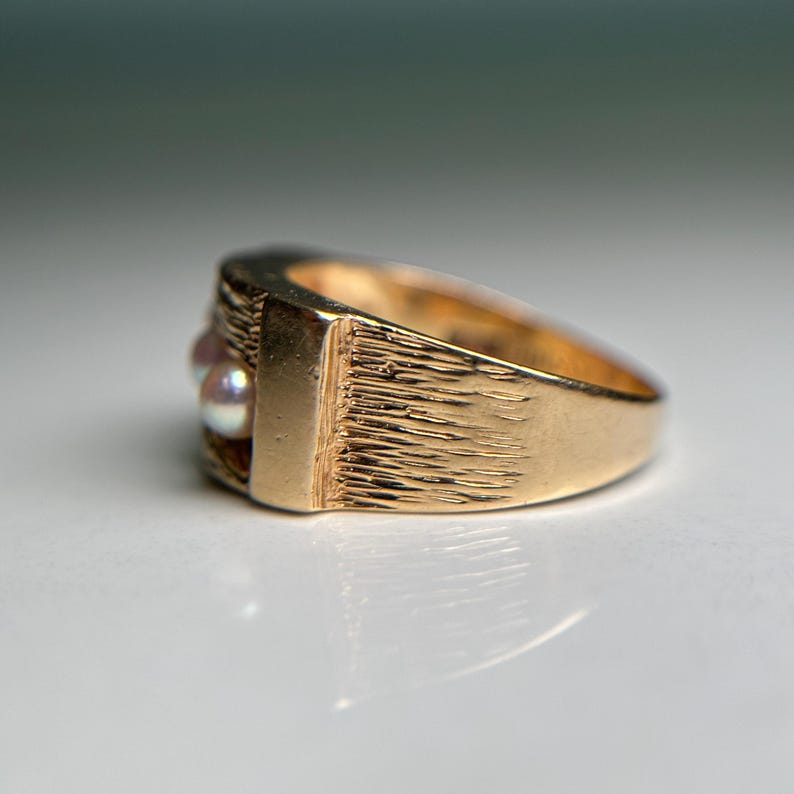 May include: A gold-colored ring with a textured band and a rectangular face. The ring features three small, round, off-white pearls. The ring is shown on a white surface.