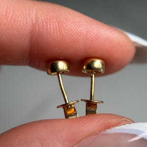 May include: A pair of small, gold-colored stud earrings. Each earring features a rounded top and a post with a small, flat backing. The earrings are held between fingers, showcasing their delicate design.