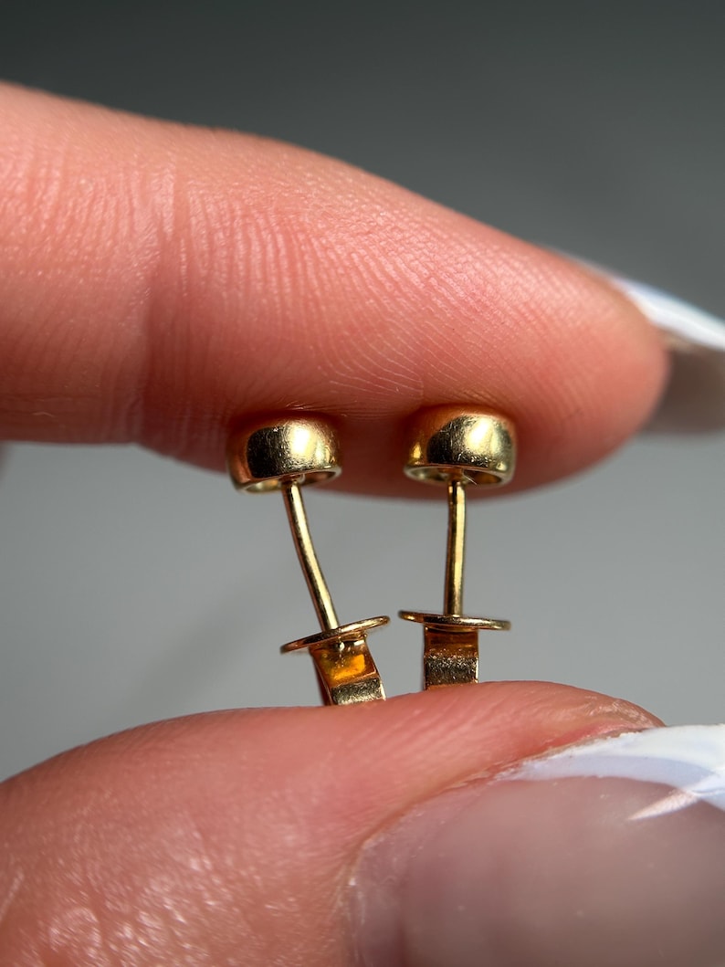May include: A pair of small, gold-colored stud earrings. Each earring features a rounded top and a post with a small, flat backing. The earrings are held between fingers, showcasing their delicate design.