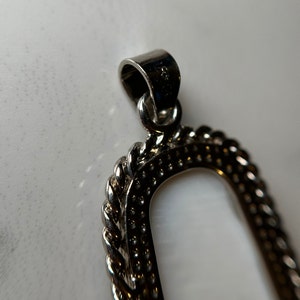 May include: Silver pendant with a rope-like border and a smooth oval-shaped center. The pendant has a loop at the top for attaching a chain.