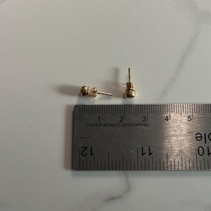 May include: A pair of small, gold-colored stud earrings with a round, possibly gemstone, center. The earrings are next to a metal ruler with centimeter markings.