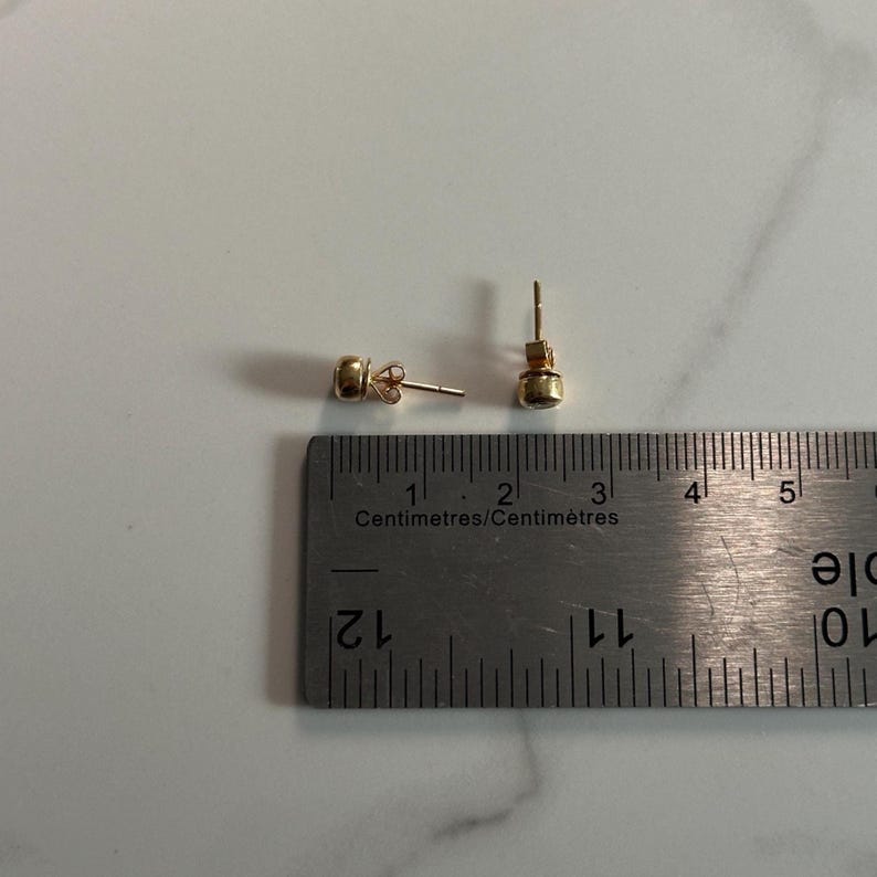 May include: A pair of small, gold-colored stud earrings with a round, possibly gemstone, center. The earrings are next to a metal ruler with centimeter markings.
