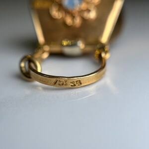 May include: Close-up of a gold-colored pendant with a loop and a small inscription. The pendant has a decorative design on the top. The loop has the inscription "750 59".