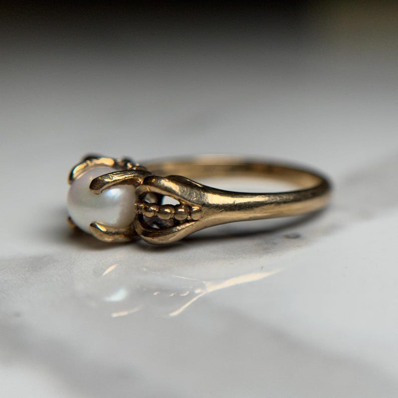 Puede incluir: Un anillo de oro con una sola perla engastada en una garra. La banda del anillo est&aacute; decorada con peque&ntilde;as cuentas.