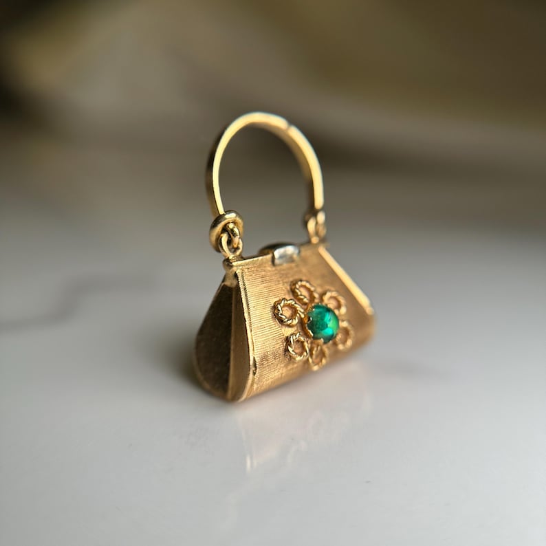 May include: A gold-colored handbag charm with a textured surface and a small handle. The charm features a flower design with a green gemstone in the center. The handbag charm is a miniature accessory.
