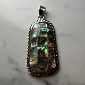 May include: A silver pendant with a rectangular abalone shell inlay. The shell is iridescent with shades of green, blue, and gold. The pendant is set in a silver frame with a rope-like border.