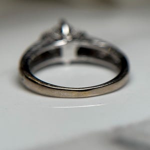 May include: A silver ring with a large center stone and smaller stones surrounding it. The ring is on a white surface.