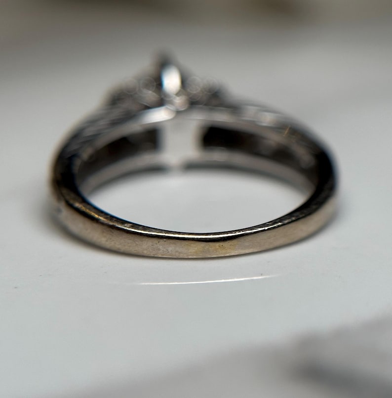 May include: A silver ring with a large center stone and smaller stones surrounding it. The ring is on a white surface.