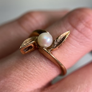 May include: A gold-toned ring featuring a central pearl and leaf-like accents. The ring's design incorporates delicate, textured details on the leaves, and the pearl has a soft, luminous quality. The ring is shown on a finger.