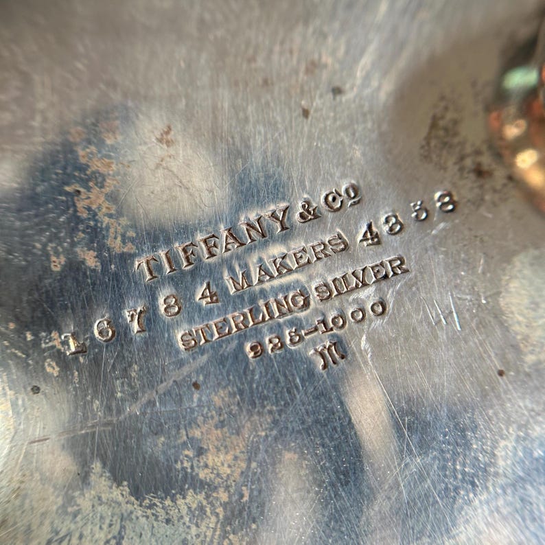May include: Close-up of a silver hallmark on a piece of silverware. The hallmark reads "Tiffany & Co. 167 8 4 Makers 483 Sterling Silver 925-1000  JM".