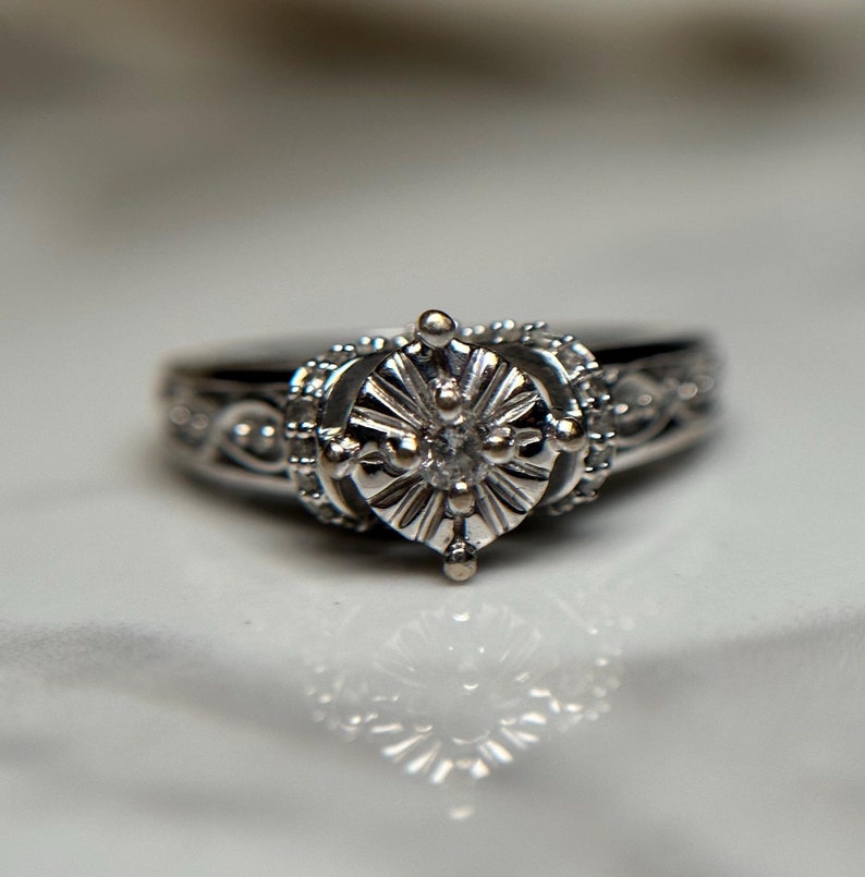 May include: A silver ring with a large, round, clear diamond set in a detailed, ornate design. The ring is set on a white surface.