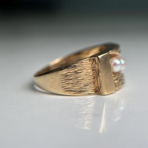 May include: A gold-colored ring with a textured band and a rectangular setting. The setting holds a small, iridescent pearl. The ring has a classic, vintage design.